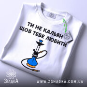 Футболка ти не кальян. Біла футболка, складена на столі, з видимим принтом на грудях. Текст 'ТИ НЕ КАЛЬЯН ЩОБ ТЕБЕ ЛЮБИТИ' та малюнок кальяну. На футболці є етикетка з зеленим шнурком. Фон світлий, з легким градієнтом.