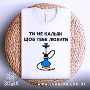 Футболка ти не кальян. Біла футболка, складена на плетеному кошику, з видимим принтом на грудях. Текст 'ТИ НЕ КАЛЬЯН ЩОБ ТЕБЕ ЛЮБИТИ' та малюнок кальяну. Фон світлий, з плетеним кошиком на передньому плані.