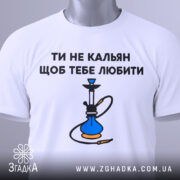 Футболка ти не кальян. Біла футболка, складена на столі, з видимим принтом на грудях. Текст 'ТИ НЕ КАЛЬЯН ЩОБ ТЕБЕ ЛЮБИТИ' та малюнок кальяну. На футболці є етикетка з зеленим шнурком. Фон світлий, з легким градієнтом.