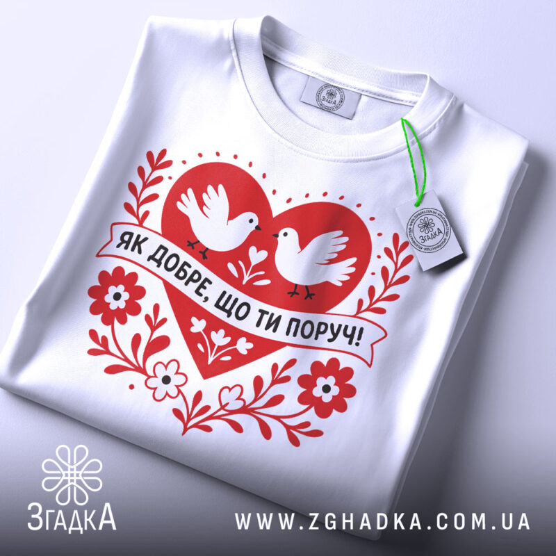 Футболка ти поруч. Білий колір, зображена зверху, на плоскій поверхні. Принт червоного серця з двома білими голубами та написом 'ЯК ДОБРЕ, ЩО ТИ ПОРУЧ!' у чорному кольорі, поряд з етикеткою зеленого кольору.