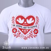 Футболка ти поруч. Білий колір, зображена зверху, на плоскій поверхні. Принт червоного серця з двома білими голубами та написом 'ЯК ДОБРЕ, ЩО ТИ ПОРУЧ!' у чорному кольорі, поряд з етикеткою зеленого кольору.