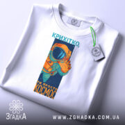 Футболка ти просто космос, біла, зображена зверху, фон світло-сірий, принт з космонавтом у оранжевому скафандрі, на футболці є етикетка з зеленим шнурком.