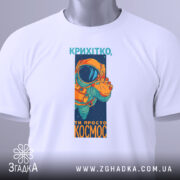 Футболка ти просто космос, біла, зображена зверху, фон світло-сірий, принт з космонавтом у оранжевому скафандрі, на футболці є етикетка з зеленим шнурком.