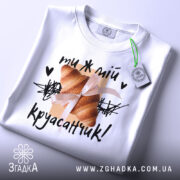 Футболка ти ж мій круасанчик. Біла футболка, складена на поверхні, з видимим коміром. На фоні світло-сірий колір. Принт з одним круасаном, обв'язаним стрічкою, та написом "ти ж мій круасанчик!" у чорному кольорі, з зеленим ярликом.