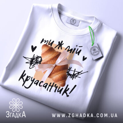 Футболка ти ж мій круасанчик. Біла футболка, складена на поверхні, з видимим коміром. На фоні світло-сірий колір. Принт з одним круасаном, обв'язаним стрічкою, та написом "ти ж мій круасанчик!" у чорному кольорі, з зеленим ярликом.