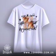 Футболка ти ж мій круасанчик. Біла футболка з коротким рукавом, зображена з фронтального ракурсу. На фоні світло-сірий колір. Принт з двома круасанами, обв'язаними стрічкою, та написом "ти ж мій круасанчик!" у чорному кольорі.