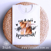 Футболка ти ж мій круасанчик. Біла футболка, складена на плетеній основі, з видимим коміром. На фоні світло-сірий колір. Принт з одним круасаном, обв'язаним стрічкою, та написом "ти ж мій круасанчик!" у чорному кольорі.