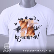 Футболка ти ж мій круасанчик. Біла футболка, складена на поверхні, з видимим коміром. На фоні світло-сірий колір. Принт з одним круасаном, обв'язаним стрічкою, та написом "ти ж мій круасанчик!" у чорному кольорі, з зеленим ярликом.