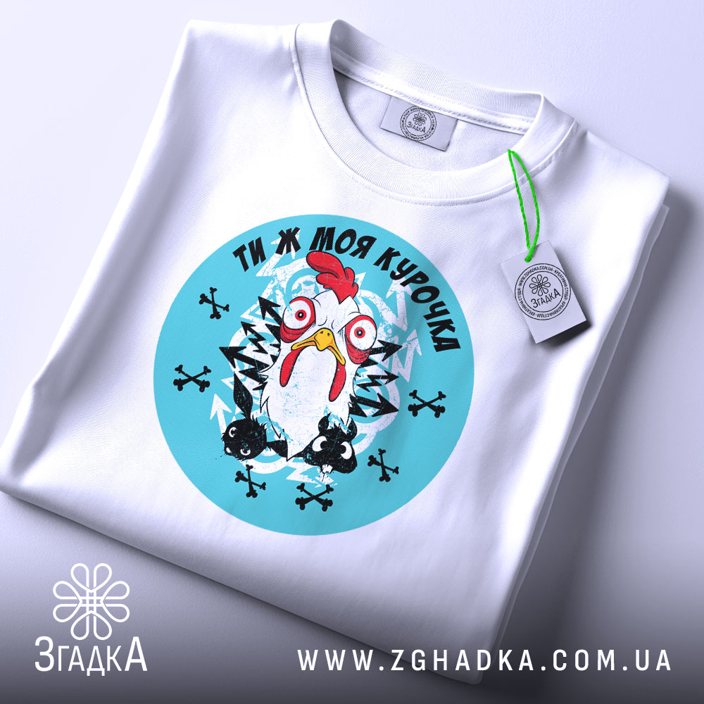 Футболка ти ж моя курочка. Біла футболка, складена на столі, з круглим принтом, що зображує курку на блакитному фоні. На футболці є етикетка з зеленим шнурком. Фон нейтральний, з легким відблиском.