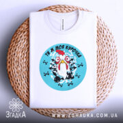 Футболка ти ж моя курочка. Біла футболка, складена на плетеному кошику, з круглим принтом курки на блакитному фоні. Принт оточений чорними хрестами. Фон світлий, з текстурою плетеного кошика.