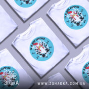 Футболка ти ж моя курочка. Біла футболка з круглим принтом, на якому зображена курка з великими очима на блакитному фоні. Принт оточений чорними хрестами та графічними елементами. Футболка знята з правого ракурсу на нейтральному фоні.