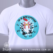 Футболка ти ж моя курочка. Біла футболка, складена на столі, з круглим принтом, що зображує курку на блакитному фоні. На футболці є етикетка з зеленим шнурком. Фон нейтральний, з легким відблиском.