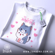 Футболка ти ж моя жопка, біла, складена на поверхні, фон світлий, принт з двома котиками, один з яких тримає іншого, навколо рожеві сердечка, на комірці є етикетка.