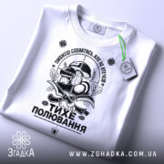 Футболка тихе полювання, білого кольору, складена на поверхні, фон світло-сірий, принт чорного кольору з елементами грибів та ножів, на футболці є зелена етикетка.