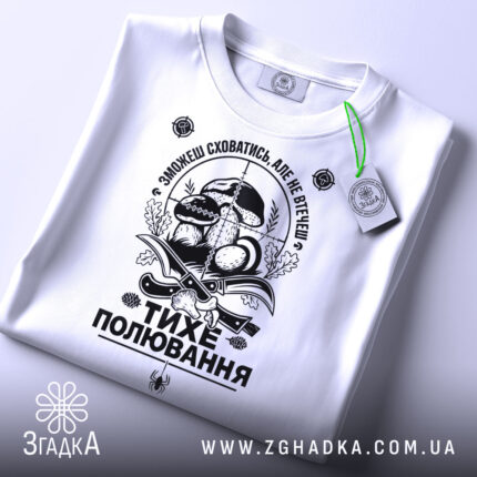 Футболка тихе полювання, білого кольору, складена на поверхні, фон світло-сірий, принт чорного кольору з елементами грибів та ножів, на футболці є зелена етикетка.