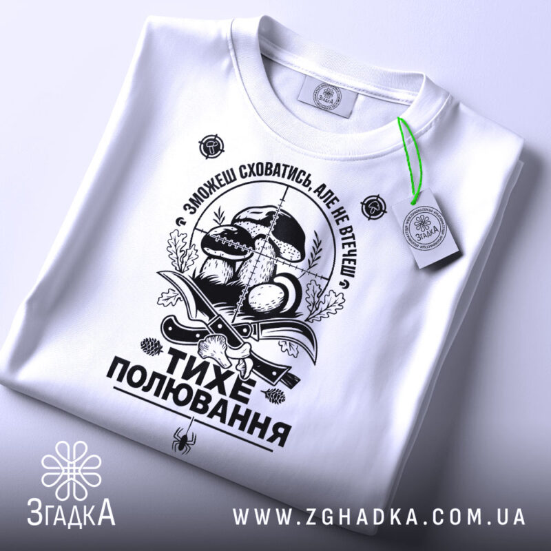 Футболка тихе полювання, білого кольору, складена на поверхні, фон світло-сірий, принт чорного кольору з елементами грибів та ножів, на футболці є зелена етикетка.