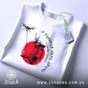 Футболка у самурая нема дрона. Біла футболка, складена на поверхні, принт самурая на червоному фоні, деталі принта чіткі, на футболці є етикетка з зеленим шнурком, фон світлий.