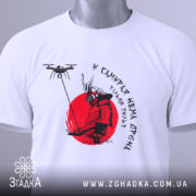 Футболка у самурая нема дрона. Біла футболка, складена на поверхні, принт самурая на червоному фоні, деталі принта чіткі, на футболці є етикетка з зеленим шнурком, фон світлий.