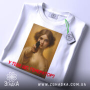 Футболка у тебе генератор. Біла футболка, складена в горизонтальному положенні, на ній принт з жінкою, що тримає пістолет, знімок з верхнього ракурсу, фон світлий, текст червоним кольором, на футболці є етикетка.