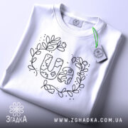 Футболка UA квіти, біла, складена на столі, з верхнього ракурсу, фон світло-сірий, принт з написом UA в оточенні квітів і листя, виконаний чорним кольором, з етикеткою зеленого кольору.