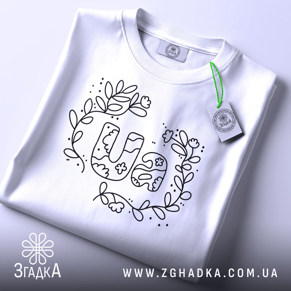 Футболка UA квіти, біла, складена на столі, з верхнього ракурсу, фон світло-сірий, принт з написом UA в оточенні квітів і листя, виконаний чорним кольором, з етикеткою зеленого кольору.