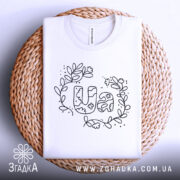 Футболка UA квіти, біла, складена на кошику, з верхнього ракурсу, фон світло-сірий, принт з написом UA в оточенні квітів і листя, виконаний чорним кольором.