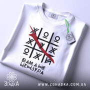 Футболка білого кольору, складена на поверхні, з принтом у вигляді гри хрестики-нулики, закресленою літерою та текстом 'Вам, а не цензура'. На футболці є етикетка з зеленим шнурком. Фон світлий, з легким градієнтом.