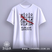 Футболка білого кольору, підвішена на вішалці, з принтом у вигляді гри хрестики-нулики, закресленою літерою та текстом 'Вам, а не цензура'. Фон світлий, з легким градієнтом.