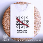 Футболка білого кольору, складена на плетеній кошику, з принтом у вигляді гри хрестики-нулики, закресленою літерою та текстом 'Вам, а не цензура'. Фон світлий, з легким градієнтом.