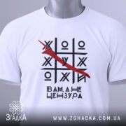Футболка білого кольору, складена на поверхні, з принтом у вигляді гри хрестики-нулики, закресленою літерою та текстом 'Вам, а не цензура'. На футболці є етикетка з зеленим шнурком. Фон світлий, з легким градієнтом.