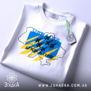 Футболка Україна авіація, біла, складена на поверхні, фон світло-сірий, принт з синім, жовтим та чорним кольорами, з контуром карти України та літаками, з біркою на комірці.
