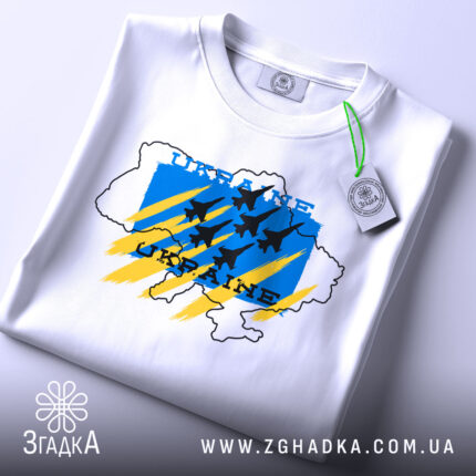 Футболка Україна авіація, біла, складена на поверхні, фон світло-сірий, принт з синім, жовтим та чорним кольорами, з контуром карти України та літаками, з біркою на комірці.