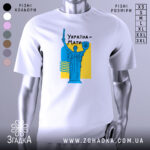 Футболка Україна Мати, біла, зображена на манекені, фон світло-сірий, принт з синьою статуєю та жовтим фоном, текст "Україна-Мати" чорним шрифтом.