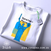 Футболка Україна Мати, біла, розкладена на поверхні, фон світло-сірий, принт з синьою статуєю та жовтим фоном, текст "Україна-Мати" чорним шрифтом, з етикеткою на комірці.