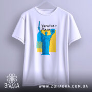 Футболка Україна Мати, біла, підвішена на вішалці, фон світло-сірий, принт з синьою статуєю та жовтим фоном, текст "Україна-Мати" чорним шрифтом.