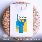 Футболка Україна Мати, біла, складена на плетеній основі, фон світло-сірий, принт з синьою статуєю та жовтим фоном, текст "Україна-Мати" чорним шрифтом.