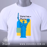 Футболка Україна Мати, біла, розкладена на поверхні, фон світло-сірий, принт з синьою статуєю та жовтим фоном, текст "Україна-Мати" чорним шрифтом, з етикеткою на комірці.