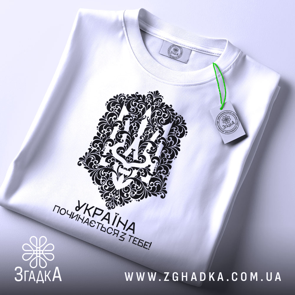 Футболка Україна починається з тебе. Біла футболка, складена на поверхні, з видимим принтом чорного кольору. На футболці видно етикетку з зеленим шнурком, що підкреслює стильний вигляд.