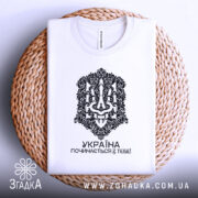 Футболка Україна починається з тебе. Біла футболка, складена на плетеній кошику, з чітким чорним принтом. Напис 'УКРАЇНА ПОЧИНАЄТЬСЯ З ТЕБЕ!' розташований під орнаментом.