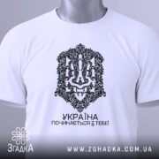 Футболка Україна починається з тебе. Біла футболка, складена на поверхні, з видимим принтом чорного кольору. На футболці видно етикетку з зеленим шнурком, що підкреслює стильний вигляд.