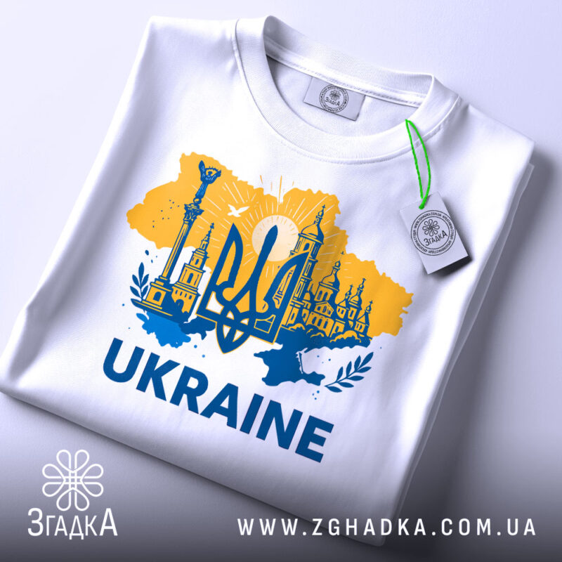 Футболка Україна тризуб, біла, зображена в плоскому вигляді, фон світло-сірий, принт жовто-блакитний з тризубом та архітектурними елементами, текст 'UKRAINE' великими буквами, на комірці є етикетка.