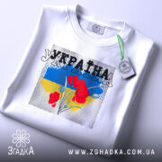 Футболка Україна білого кольору, складена на столі, з принтом червоних квітів на фоні синьо-жовтого кольору, ракурс зверху, видно етикетку з зеленим шнурком.