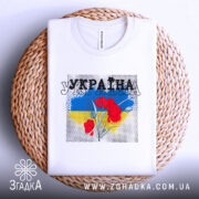 Футболка Україна білого кольору, складена на плетеному кошику, з принтом червоних квітів на фоні синьо-жовтого кольору, ракурс зверху, фон білий.