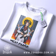 Футболка українка незламна, біла, зображена зверху, фон світло-сірий, принт з жінкою в національному вбранні з автоматом, на футболці є етикетка з зеленим шнурком.