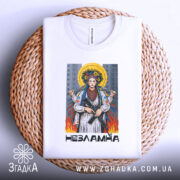 Футболка українка незламна, біла, зображена на плетеній кошику, фон світло-сірий, принт з жінкою в національному вбранні з автоматом, оточена вогнем і містом на задньому плані.