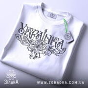 Футболка українка білого кольору, складена на поверхні, зображена зверху, фон світло-сірий, принт з написом 'Україна' в стилізованому шрифті, на футболці підвішена етикетка з зеленим шнурком.