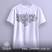 Футболка українка білого кольору, зображена на вішалці з фронтального ракурсу, фон світло-сірий, принт з написом 'Україна' в стилізованому шрифті з витонченими візерунками.