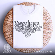 Футболка українка білого кольору, складена на плетеній кошику, зображена зверху, фон світло-сірий, принт з написом 'Україна' в стилізованому шрифті, видима етикетка на комірці.