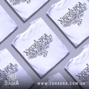 Футболка українка білого кольору, зображена з фронтального ракурсу на манекені, фон світло-сірий, принт з написом 'Україна' в стилізованому шрифті з витонченими візерунками.