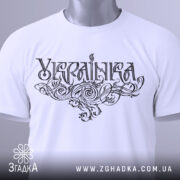 Футболка українка білого кольору, складена на поверхні, зображена зверху, фон світло-сірий, принт з написом 'Україна' в стилізованому шрифті, на футболці підвішена етикетка з зеленим шнурком.